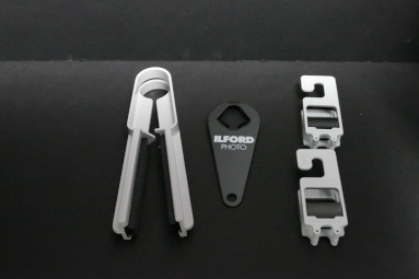 A set of photographic tools including a gray clipper, a black ILFORD Photo tag, and two gray film holders, all arranged on a dark surface.